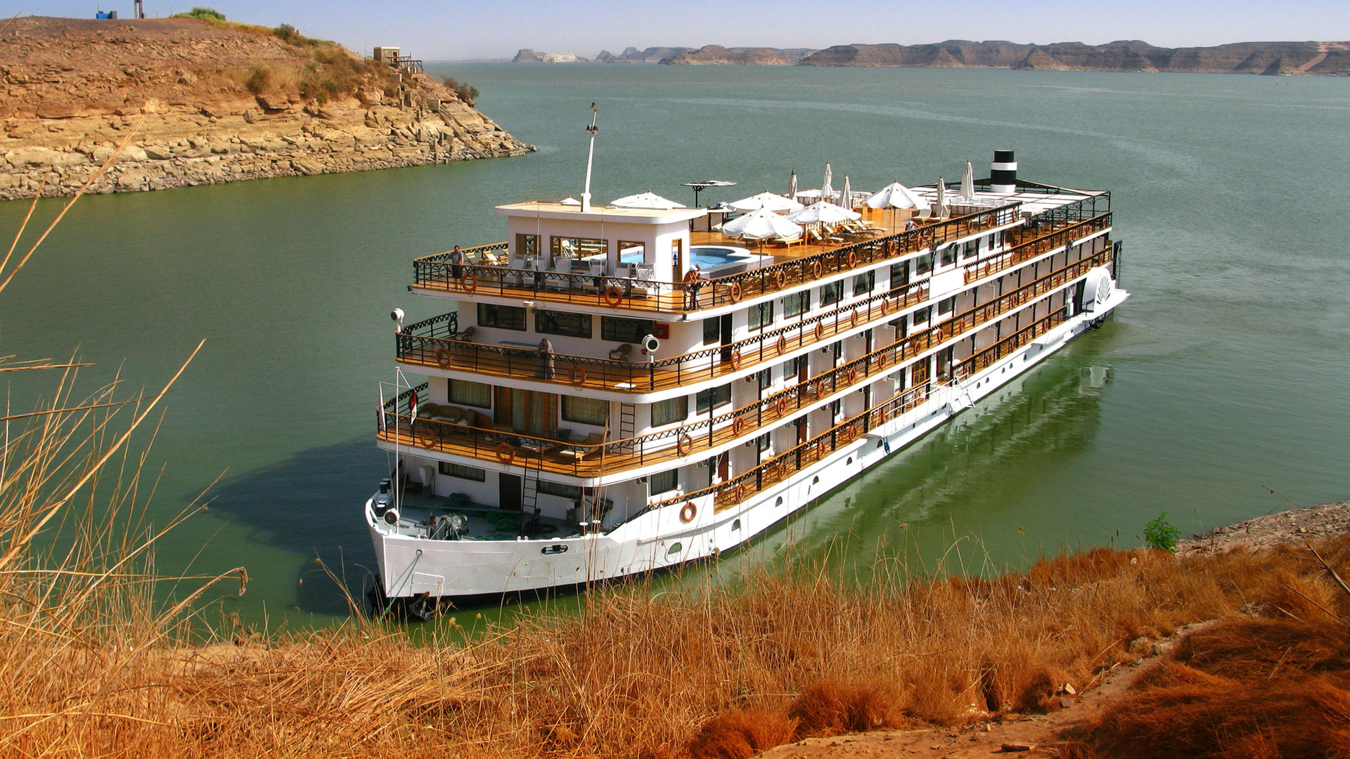 Cruise ship with wooden decks moored against a rocky desert shore